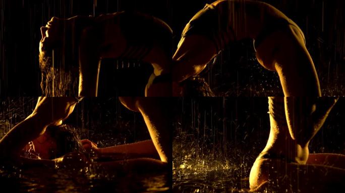 剪影被雨淋湿的苗条女人在黑暗的工作室里练习瑜伽体式。性感的身体覆盖着在工作室黄光下闪闪发光的水滴。黑