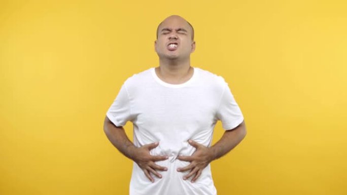 疲倦的不健康的秃头男人看着相机把手放在肚子上肚子疼穿着白色t恤孤立在黄色背景上。4k分辨率。