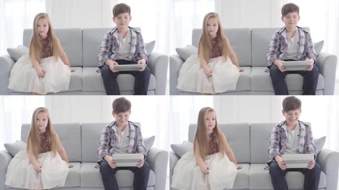 两个白人孩子坐在沙发上的肖像。男孩在平板电脑屏幕上看东西，女孩穿着漂亮的衣服看着远处。初恋，休闲。