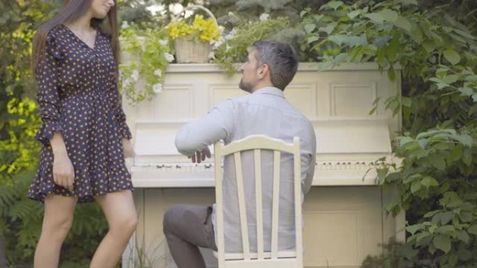灰发的成年男子被美女分散了弹钢琴的注意力。快乐浪漫的高加索钢琴家在夏日花园与女友交谈。休闲概念
