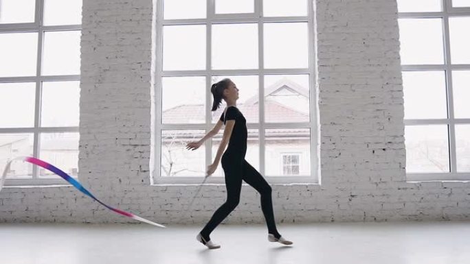 艺术体操的训练-十几岁的女孩在白色工作室或体操学校的背景大窗户上用彩带进行体操练习。穿着黑色身体的苗