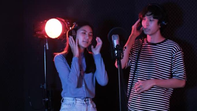 年轻的亚洲有吸引力的音乐家快乐地一起唱歌，在现代录音棚录制新歌。两个年轻人在黑暗背景下的工作室里一起