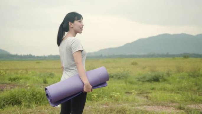 迷人的亚洲女人早上站在美丽的山景里拿着瑜伽垫。