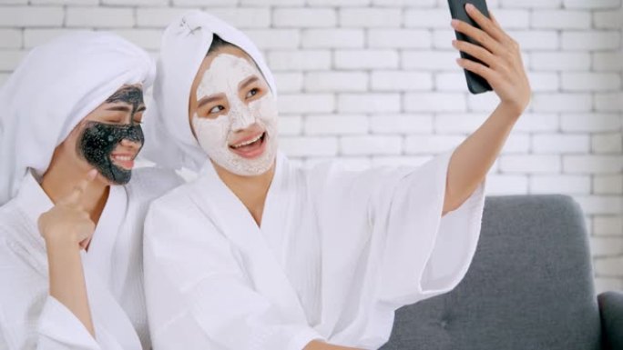 两个穿着白色浴袍戴着面膜的快乐亚洲女孩在客厅一起用智能手机拍照。