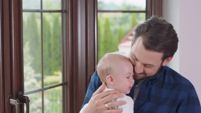 留着胡须，留着胡须的父亲抱着他可爱的婴儿，呆在窗户旁边，腿上微微波动，微笑着，将脸颊压在额头上，亲吻