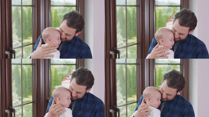 留着胡须，留着胡须的父亲抱着他可爱的婴儿，呆在窗户旁边，腿上微微波动，微笑着，将脸颊压在额头上，亲吻