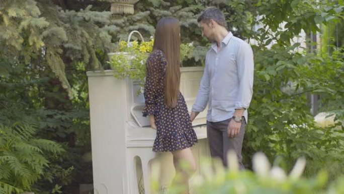 可爱的高加索夫妇站在复古钢琴旁边聊天。快乐的成年男子和年轻女子在夏日聊天并微笑。女友和男朋友在户外约