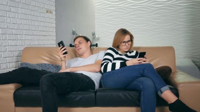 迷人的年轻夫妇坐在沙发上背对背，痴迷于智能手机。与手机相视对方为陌生人的夫妻，沟通问题和社交网络成瘾