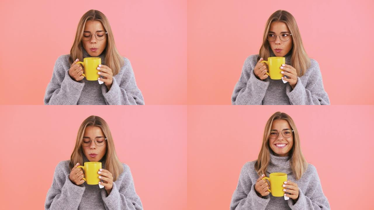 年轻的女士拿着一杯黄色的茶，吹凉，微笑着在粉红色的工作室背景下摆姿势