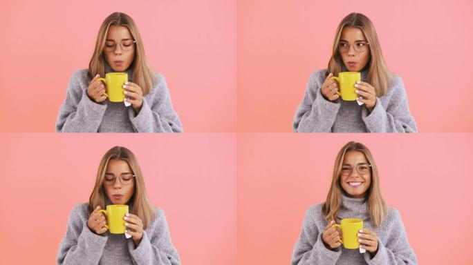 年轻的女士拿着一杯黄色的茶，吹凉，微笑着在粉红色的工作室背景下摆姿势