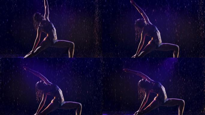 年轻女子在工作室蓝光和水雨下进行瑜伽练习。水滴中的女性身体。在黑色背景的工作室里拍摄的照片。慢动作