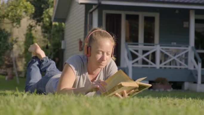 戴着耳机的女人躺在草坪上看书。