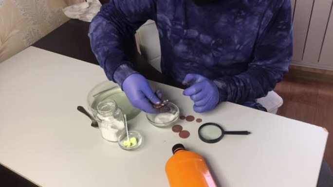 一个戴着口罩和橡胶手套的人用苏打水和水稀粥擦拭铜币。附近有磷酸和硫酸软膏清洗铜币