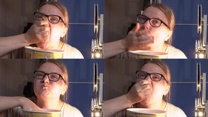 戴着眼镜的年轻有趣的女人吃爆米花，贪婪地将大部分塞入嘴里。附近有一台工作的老式电影放映机。愉快家庭影