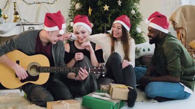 一个穿着圣诞老人帽子的家伙在圣诞树附近弹吉他。朋友在阁楼房间庆祝圣诞节，跳舞唱歌，玩得开心