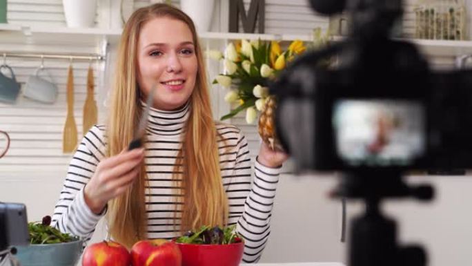 食物博主在厨房工作室烹饪新鲜的水果素食沙拉，在视频频道的相机上拍摄教程。女性影响者手持苹果、菠萝，谈