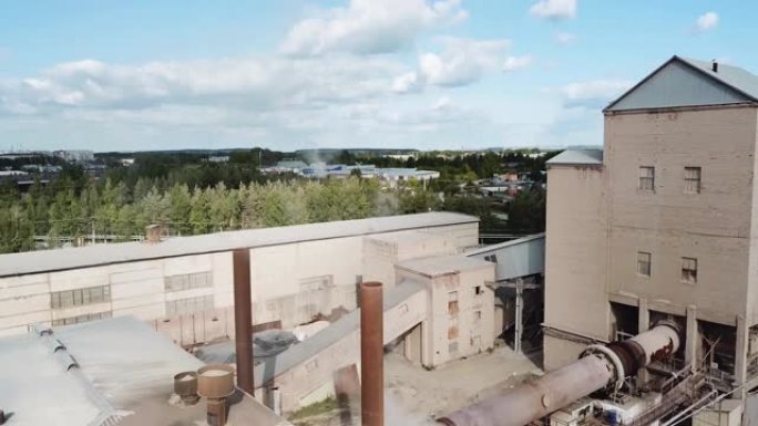 一个有吸烟烟囱的小工厂的鸟瞰图。库存镜头。在蓝色多云的天空背景下，工厂上空被绿树环绕，工业工程概念