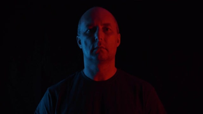 蓝色和红色照明的黑色背景上的肖像成年男子。黑暗工作室背景下的男性脸看着镜头。