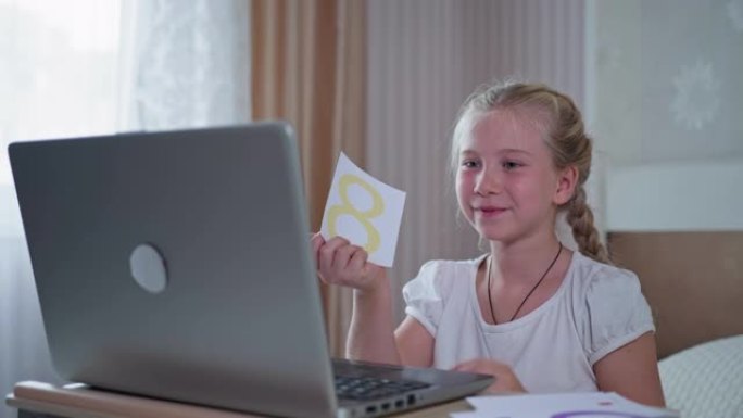 远程教育在大流行期间，可爱的小女孩坐在家里拿着笔记本电脑，在学校在线课程上给老师看卡片