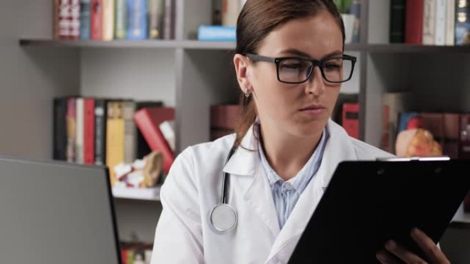 医生将数据输入计算机。办公室工作场所穿着白大褂的专注女医生将纸质病历卡中的数据输入计算机内存