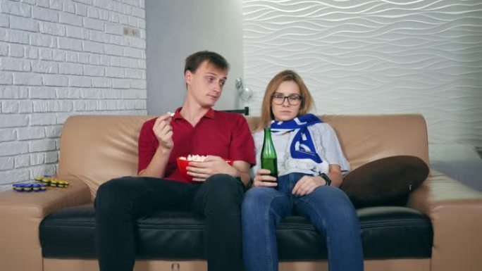 一对年轻男女坐在电视机前，一边吃着爆米花，一边看无聊的体育比赛。女人的姿势表示不喜欢。概念:运动、休