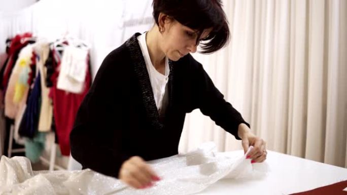 女裁缝，熟练的工人从白色闪闪发光的织物上取别针。白布，婚纱和裁缝钉在桌子上的材料。概念缝纫和手工艺