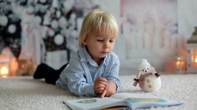 金发蹒跚学步的孩子，男孩，在家看书，躺在地板上，拿着玩具
