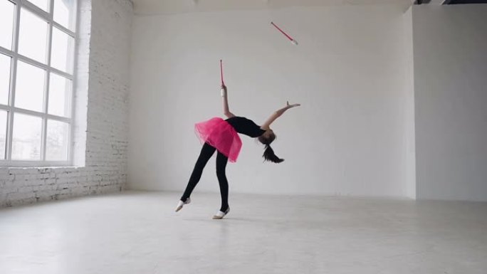 可爱的体操运动员在窗户背景的白色工作室里用狼牙棒在腿上打圈。体操女孩穿着黑色紧身衣和粉色裙子