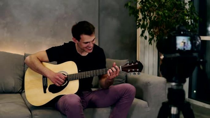 穿着黑色t恤的英俊白人男子在家里坐在沙发上时，制作有关乐器的视频博客或为追随者录制歌曲。在前景上记录