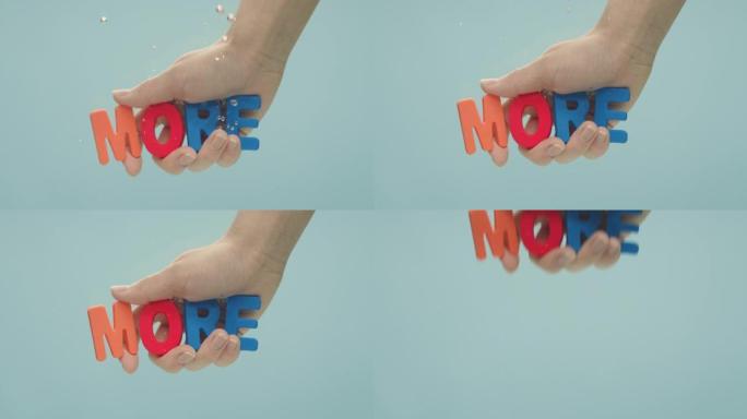 手将 “更多” 一词浸入水中，该词由彩色塑料字母拼写而成。