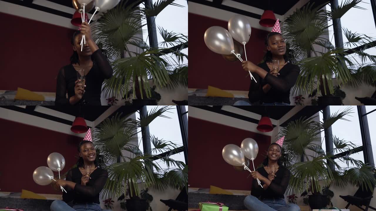 戴着帽子的迷人非洲女孩，一只手拿着香槟玻璃，另一只手拿着气球坐在红色房间的地板上