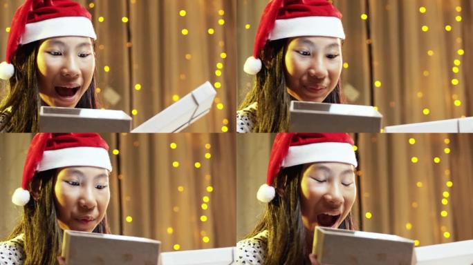 快乐的亚洲女孩戴着圣诞老人的帽子，在家打开带圣诞灯的礼品盒。