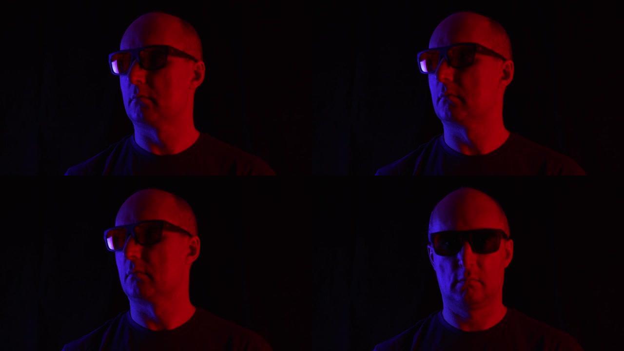 黑眼镜的秃头男人在蓝色和红色的灯光下。穿着墨镜的肖像成年男子在黑暗的工作室里看着相机，背景上有蓝色和