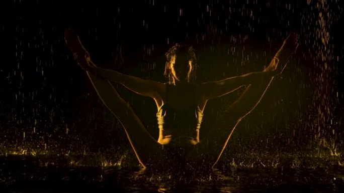 一个年轻的女人在雨中黑暗的工作室里保持平衡，双腿分开。在工作室的黑色背景上，水滴和黄色灯光映衬着苗条