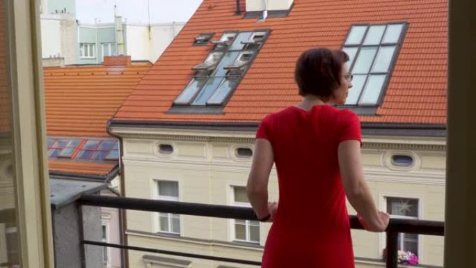 旅行中的女性肖像。穿着红色连衣裙的美丽迷人的女人去卧室的阳台，看着早晨的城市，放松。现代公寓的内部。