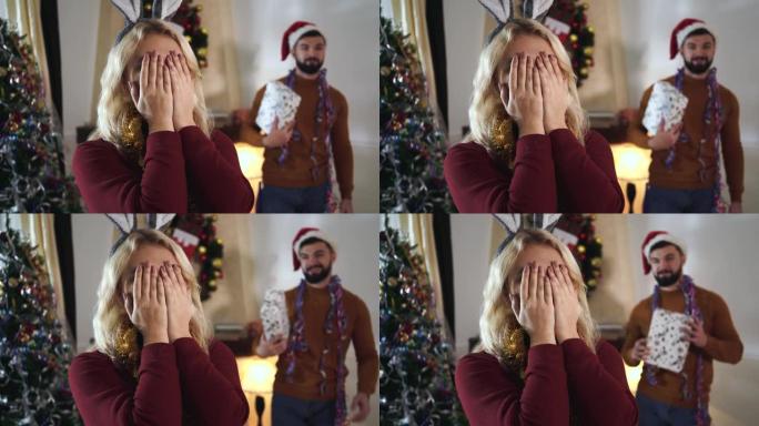 金发碧眼的高加索女人用手闭上眼睛的头像，戴着圣诞帽的大胡子男人站在礼品盒的背景下。男女朋友在家一起庆
