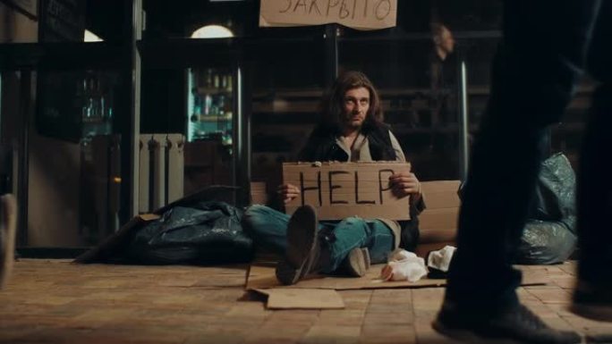 路人走在乞求无家可归的人附近，在夜间城市街道4K上有标志帮助。