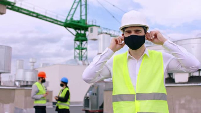 建筑工地冠状病毒的概念屋顶上有安全帽的商人脱下防护面罩背景上的其他建筑工人戴着防护面罩在背景上讨论大