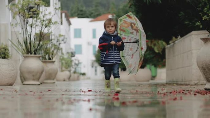 雨中，一个穿着橡胶靴的小男孩拿着雨伞穿过水坑