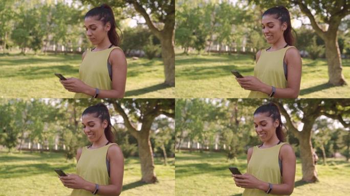 一位穿着运动服的美丽健康微笑的年轻女子的肖像站在公园里用智能手机发短信