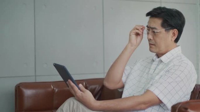 坐在房子里的亚洲老人用平板电脑看书，
由于长期使用视力，高级男子患有视力乏力。眼睛健康问题