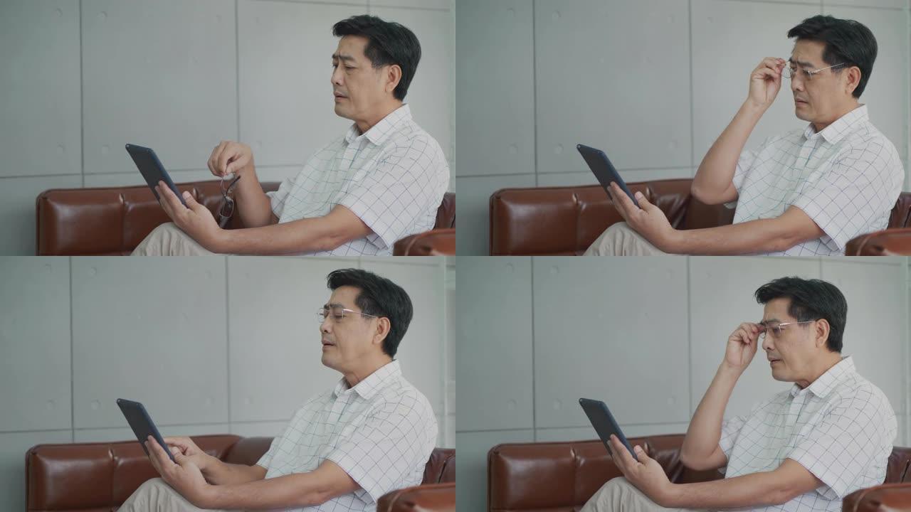 坐在房子里的亚洲老人用平板电脑看书，
由于长期使用视力，高级男子患有视力乏力。眼睛健康问题