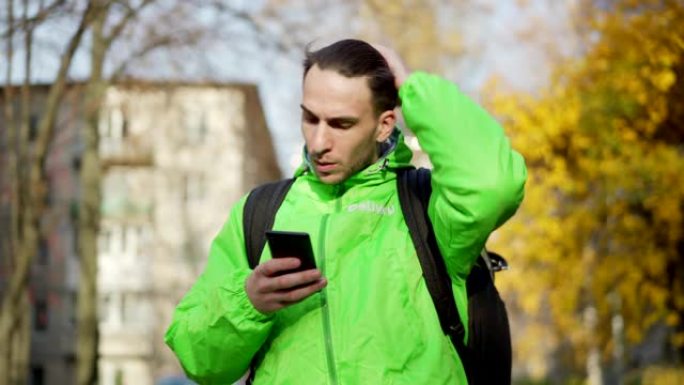 穿着绿色制服的年轻食品快递员的腰部拍摄，使用智能手机上的移动导航应用程序在秋天的街道上寻找方向