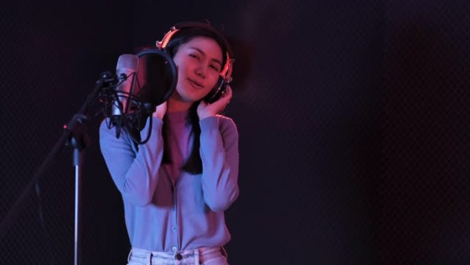 年轻的亚洲女性迷人的音乐家快乐唱歌在现代录音棚录制新歌。美丽的女性在黑暗背景下的工作室麦克风附近工作