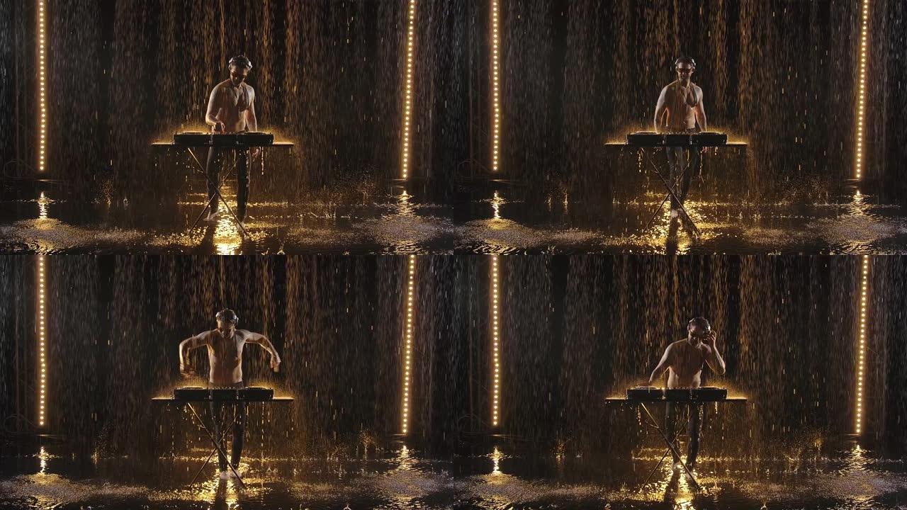 一个男性裸胸DJ在转盘上播放电子舞曲，并在黑暗的工作室里享受雨水。在慢动作的黑暗背景下拍摄视频