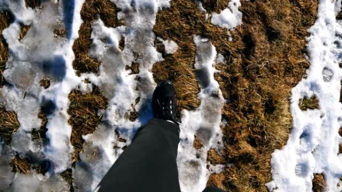 穿着靴子的男性脚的观点穿过雪域。早春时，年轻的徒步旅行者的腿沿着雪草地走。寒假或假期的概念。关闭慢动