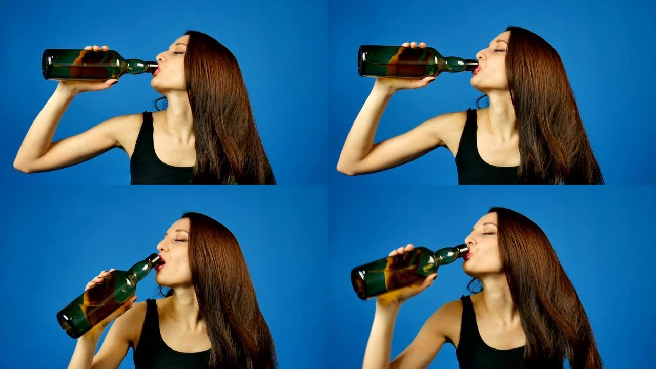 年轻的黑发女人正在工作室里喝蓝色背景的深绿色瓶子里的葡萄酒