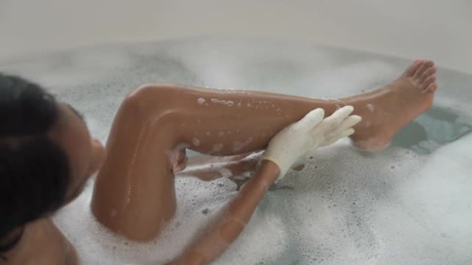 躺在泡泡浴中的非洲裔美国妇女的后视图用去角质手套洗自己。水疗和护肤概念