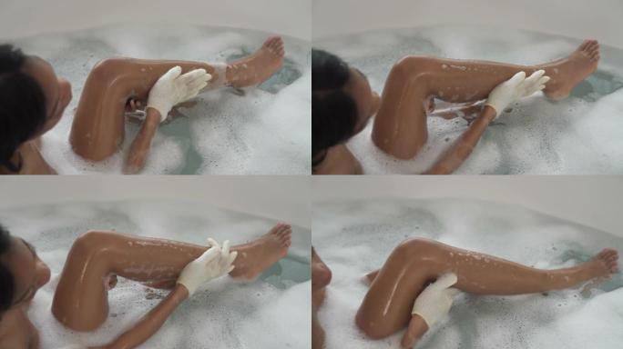 躺在泡泡浴中的非洲裔美国妇女的后视图用去角质手套洗自己。水疗和护肤概念
