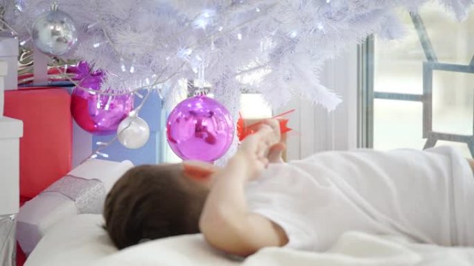 视频的小孩躺在圣诞树下，触摸球，看着他的倒影在粉红色的玻璃球。